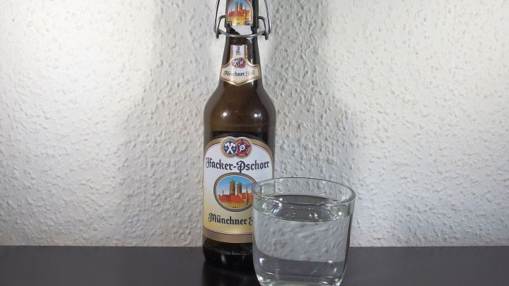 Eine Bierflasche steht neben einem Glas Wasser auf einem Tisch vor einer hellen Wand.