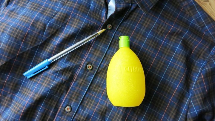 Ein gelbes Zitronensaftfl&auml;schchen liegt auf einem karierten Hemd neben einem blauen Kugelschreiber, bereit zum Fleckenentfernen.