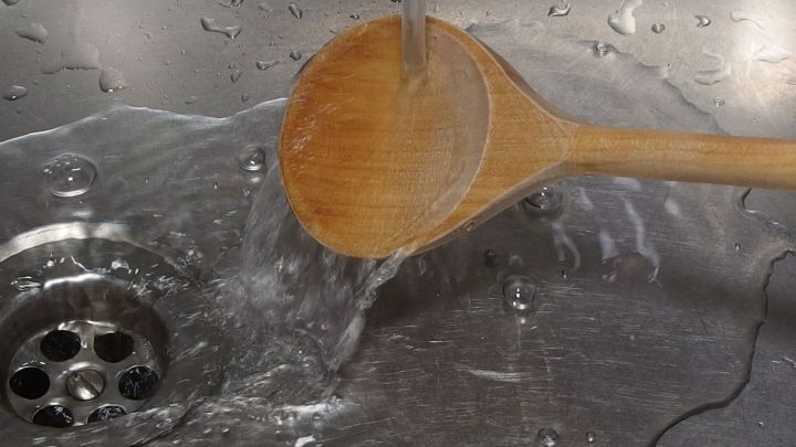Ein Holzl&ouml;ffel wird unter flie&szlig;endem Wasser gewaschen, w&auml;hrend er in einem Sp&uuml;lbecken mit Abflussl&ouml;chern liegt.
