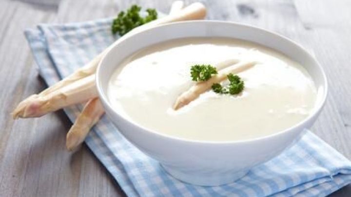 Eine Sch&uuml;ssel Spargelcremesuppe steht auf einem blau karierten Tuch, garniert mit Petersilie, daneben liegen frische Spargelstangen.