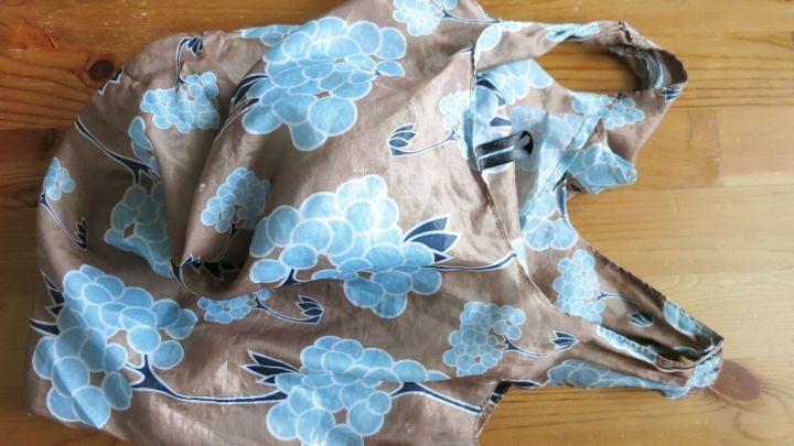 Ein faltbarer Einkaufsbeutel liegt auf einem Holztisch und pr&auml;sentiert ein florales Design in Blau und Beige.