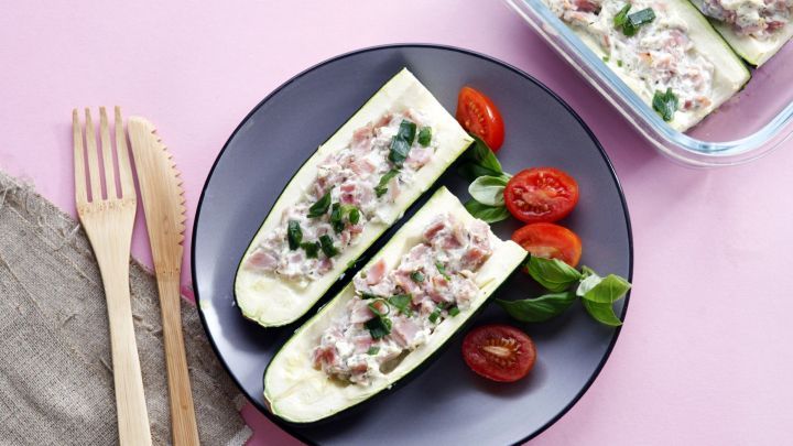 Gef&uuml;llte Zucchini liegen auf einem Teller, umgeben von Kirschtomaten und frischem Basilikum auf einem rosa Hintergrund.