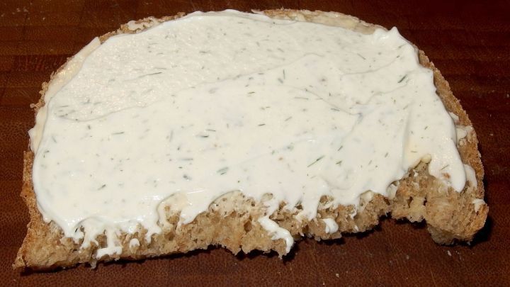 Ein St&uuml;ck Brot ist mit einer cremigen Sahne-K&auml;se-Creme bestrichen, die glatt und leicht verl&auml;uft, auf einem Holzbrett.