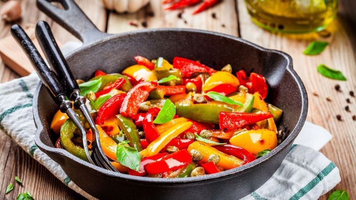 Bunte Paprika, zubereitet in einer gusseisernen Pfanne, umgeben von frischen Kr&auml;utern und Oliven&ouml;l auf einem Holztisch.