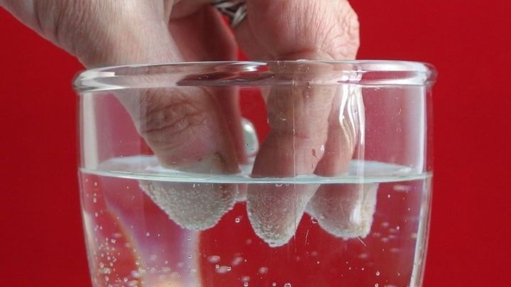Eine Hand taucht in ein Glas Wasser, um die Fingern&auml;gel zu s&auml;ubern, w&auml;hrend der Hintergrund rot ist.