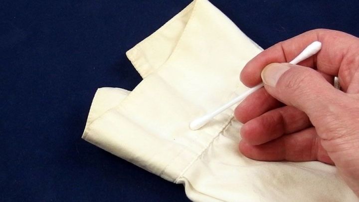 Eine Hand benutzt ein Wattest&auml;bchen, um Flecken auf einem wei&szlig;en Textil auf einem dunklen Hintergrund zu behandeln.