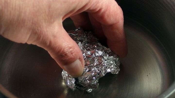 Eine Hand greift in einen Topf mit Wasser, w&auml;hrend sie ein St&uuml;ck Alufolie zum Scheuern verwendet.