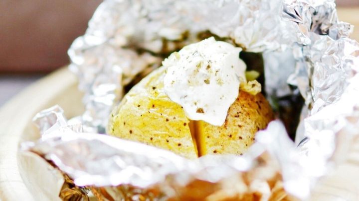 Eine Folienkartoffel mit Sour Cream liegt auf einem Brett, umgeben von Alufolie, bereit zum Servieren.