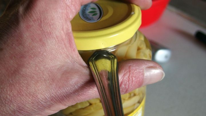 Eine Hand h&auml;lt einen Glasdeckel mit einem L&ouml;ffel, um ihn leicht zu &ouml;ffnen, w&auml;hrend er auf einer Tischoberfl&auml;che liegt.