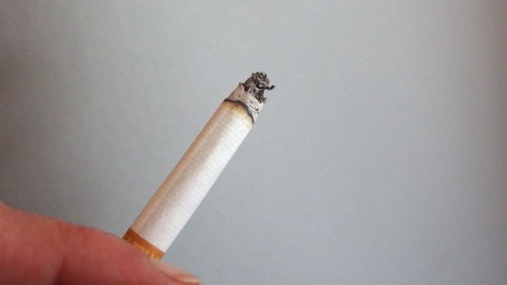 Eine brennende Zigarette wird von einer Hand gehalten, w&auml;hrend der Rauch sichtbar aufsteigt. Der Hintergrund ist neutral.