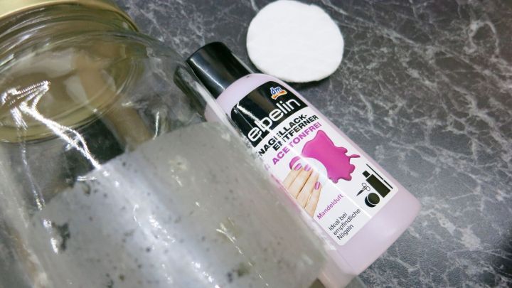 Ein Fl&auml;schchen Nagellackentferner liegt neben einem Wattepad auf einer dunklen, marmorierten Oberfl&auml;che.