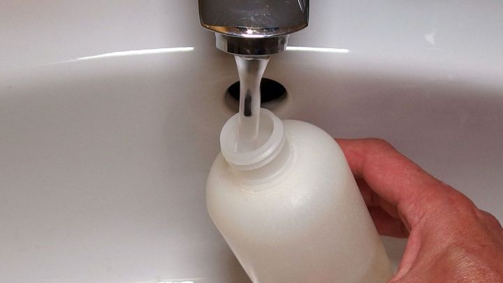 Eine Hand h&auml;lt eine Flasche Duschgel, w&auml;hrend Wasser aus dem Wasserhahn hineinl&auml;uft, um Reste nicht zu verschwenden.