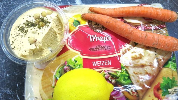 Ein Wrap mit frischem Gem&uuml;se liegt neben Hummus, einer Zitrone und Karotten auf einer Tischoberfl&auml;che.