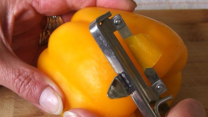 Eine Hand sch&auml;lt eine gelbe Paprika mit einem Sch&auml;ler auf einem Holzbrett in einer hellen K&uuml;chenumgebung.