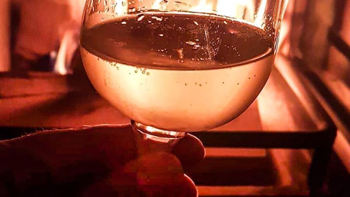 Ein Weinglas mit sprudelndem Getr&auml;nk wird von einer Hand gehalten, w&auml;hrend Flammen im Hintergrund leuchten.