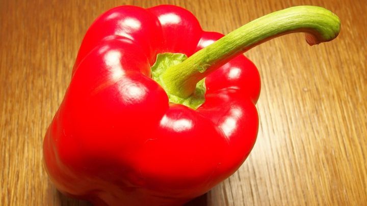 Eine rote Paprikaschote liegt auf einem Holztisch, bereit zum Sch&auml;len und Zubereiten f&uuml;r ein Gericht.