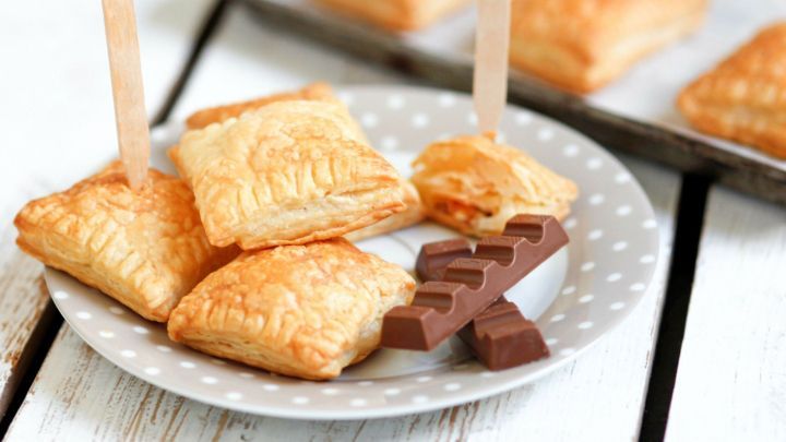 Kleine Bl&auml;tterteigtaschen mit einem Holzstab stehen auf einem Teller, neben einem St&uuml;ck Kinderschokolade auf einem Tisch.