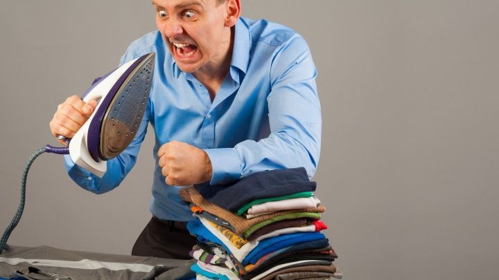 Mann h&auml;lt ein B&uuml;geleisen ver&auml;rgert hoch, neben ihm ein Stapel Kleidung auf einem B&uuml;gelbrett im hellgrauen Hintergrund.