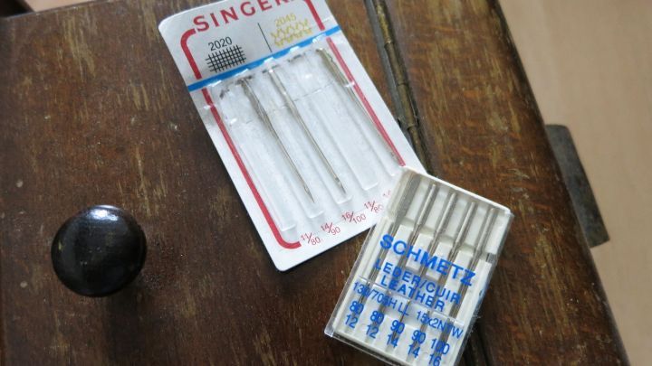 N&auml;hmaschinennadeln liegen auf einem Holztisch, umgeben von einer dunklen Schublade und einem runden Knopf.