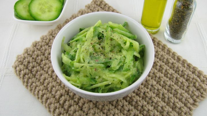 Eine Sch&uuml;ssel mit gr&uuml;nem Gurkensalat steht auf einer handgekn&uuml;pften Unterlage, umgeben von Oliven&ouml;l und Gew&uuml;rzen.