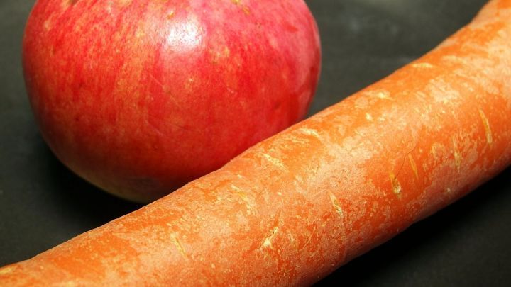 Eine frische Karotte liegt neben einem roten Apfel auf einem schwarzen Teller, ideal f&uuml;r einen gesunden Snack.