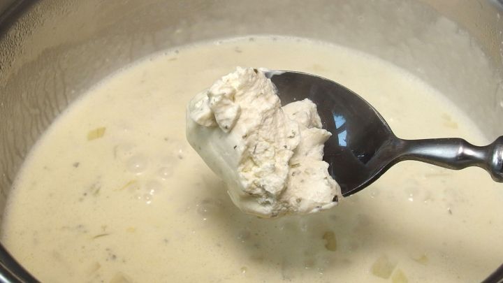 Eine L&ouml;ffel mit Frischk&auml;se wird aus einer cremigen So&szlig;e gehoben, die in einem Topf auf einem Herd k&ouml;chelt.