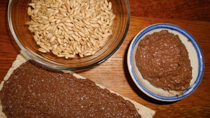 Eine cremige Schoko-Hafer-Creme liegt auf einer Brotoberfl&auml;che, daneben sind Haferflocken und eine kleine Sch&uuml;ssel mit Creme.