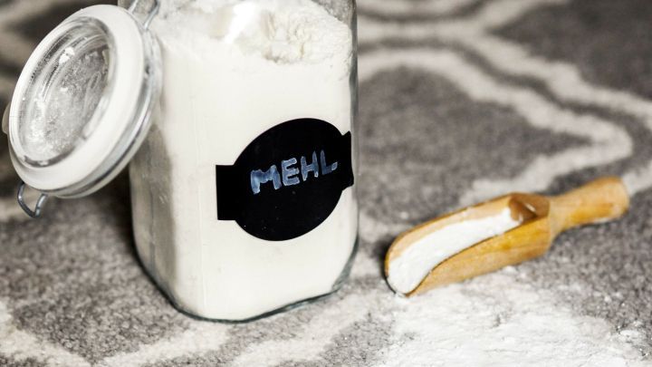 Ein Glas mit Mehl steht auf einem Teppich, w&auml;hrend ein Holzl&ouml;ffel daneben liegt, und etwas Mehl versch&uuml;ttet wurde.