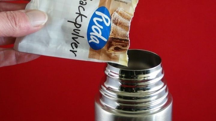 Eine Hand h&auml;lt ein P&auml;ckchen Backpulver &uuml;ber eine Thermoskanne, bereit, es hineinzusch&uuml;tten, vor rotem Hintergrund.