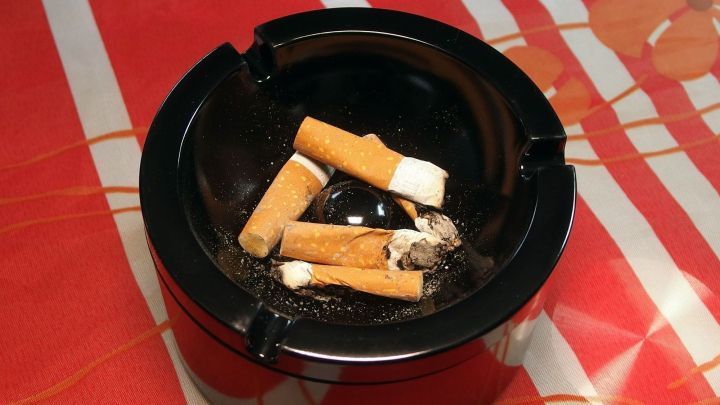 Ein schwarzer Aschenbecher steht mit vielen Zigarettenstummeln auf einem rot karierten Tisch, umgeben von Wohnungseinrichtung.