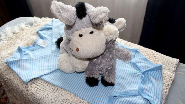 Ein Kuscheltier in Form eines Eies, umgeben von blauer Babykleidung auf einem Bett f&uuml;hrt frischgebackene Mamas in eine liebevolle Atmosph&auml;re.