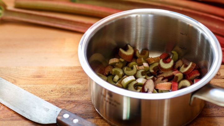 Ein Topf mit geschnittenem Rhabarber und anderen Zutaten steht auf einem Holzbrett, neben einem K&uuml;chenmesser.