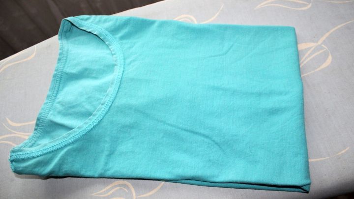 Ein frisch gefaltetes, t&uuml;rkisfarbenes T-Shirt liegt ordentlich auf einem hellen Tisch und wartet darauf, getragen oder gewaschen zu werden.