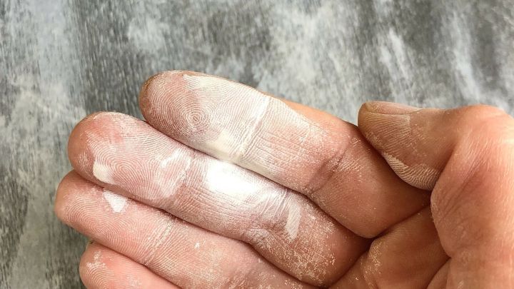 Die Hand hält eine lackierte Oberfläche mit abblätternder Farbe. Der Kontext zeigt einen grauen Untergrund, der verschmutzt ist.
