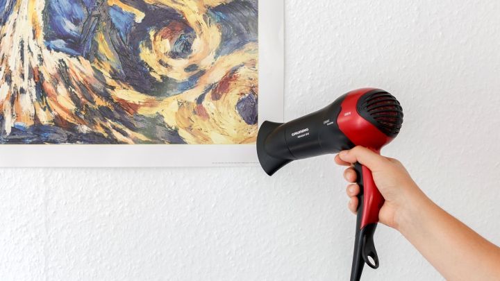 Ein F&ouml;hn bl&auml;st warme Luft auf ein an der Wand h&auml;ngendes abstraktes Poster, das anscheinend entfernt werden soll.