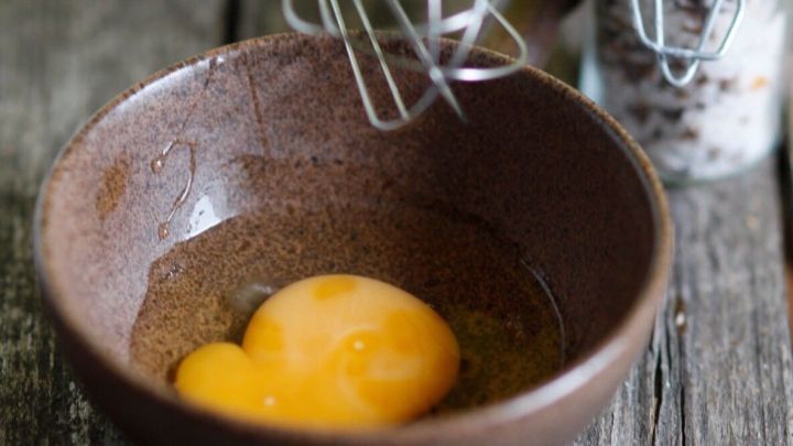 Ein gelbes Eigelb liegt in einer dunklen Sch&uuml;ssel, w&auml;hrend ein Schneebesen dar&uuml;ber schwebt, um eine farbenfrohe Zubereitung vorzubereiten.