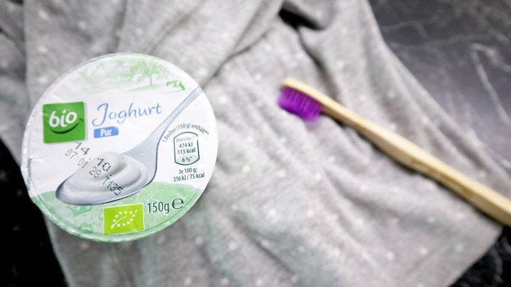 Ein Joghurtbecher liegt auf einem grauen Tuch, w&auml;hrend eine Zahnb&uuml;rste daneben f&uuml;r das Ausb&uuml;rsten der Flecken bereitsteht.
