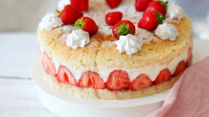 Ein luftiger Bisquitboden ist mit frischen Erdbeeren und Sahne dekoriert und steht auf einer wei&szlig;en Tortenplatte.