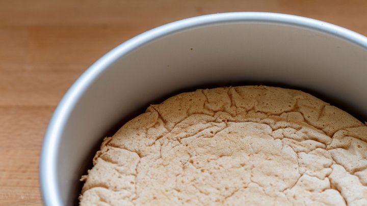 Ein Tortenboden liegt in einer runden Backform, leicht aufgegangen und mit Rissen, auf einer Holzoberfl&auml;che.