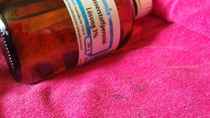 Eine Flasche Wasserstoffperoxid liegt auf einem pinken Tuch, bereit zur Anwendung gegen Blaubeerflecken.