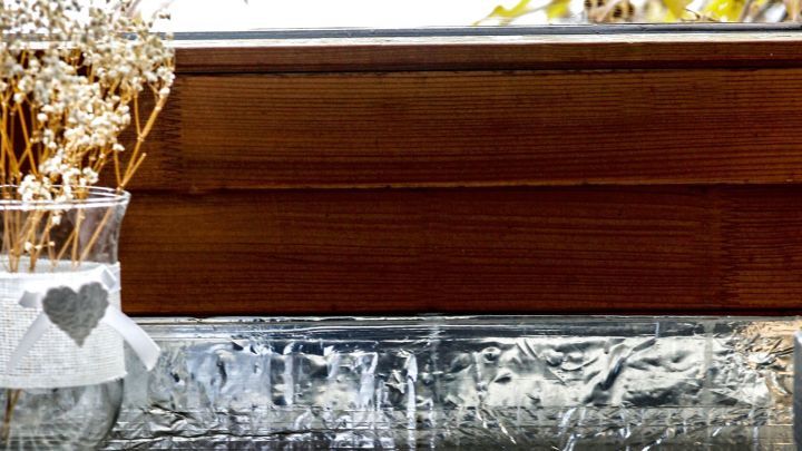 Eine Glasvase mit Trockenblumen steht auf einer Alufolie-bedeckten Fensterbank, w&auml;hrend die Bl&auml;tter drau&szlig;en im Wind wehen.