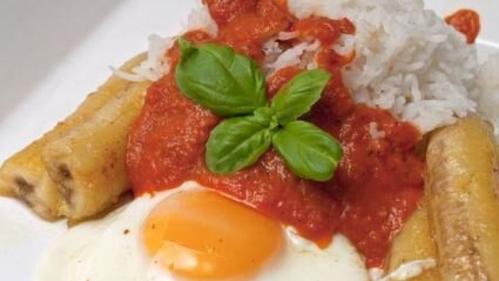 Reis mit einem Spiegelei und gebratenen Kochbananen, garniert mit Tomatensauce und Basilikum auf einem Teller angerichtet.