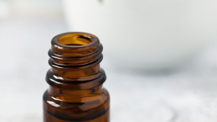 Ein braunes Fl&auml;schchen mit K&uuml;mmel&ouml;l steht auf einer hellen Oberfl&auml;che, w&auml;hrend im Hintergrund ein M&ouml;rser mit frischen Kr&auml;utern zu sehen ist.