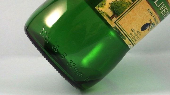 Eine leere gr&uuml;ne Oliven&ouml;lflasche steht schr&auml;g auf einer neutralen Oberfl&auml;che und zeigt das Etikett mit Olivenmotiven.