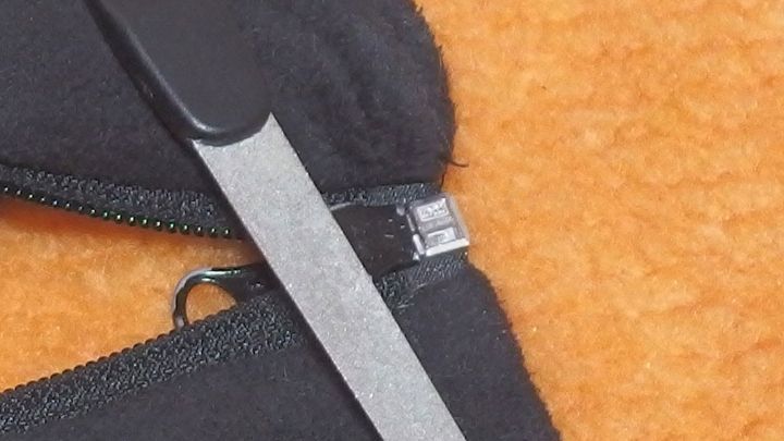 Ein ge&ouml;ffneter, schwarzer Rei&szlig;verschluss wird mit einer kleinen, silbernen Feile bearbeitet, auf einem orangefarbenen Hintergrund.