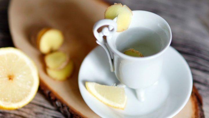 Eine helle Tasse mit Ingwertee steht auf einem Holzbrett, umgeben von Zitronenscheiben und Ingwerst&uuml;cken.