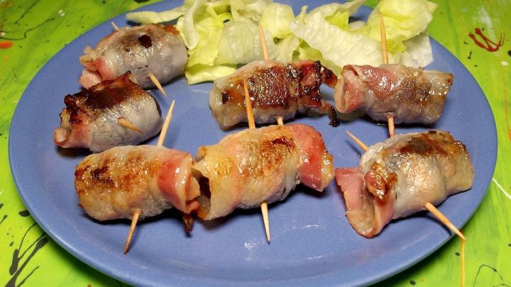 Saftige Pflaumen sind in knusprigem Speck eingewickelt und sch&ouml;n angerichtet auf einem blauen Teller, umgeben von Salat.