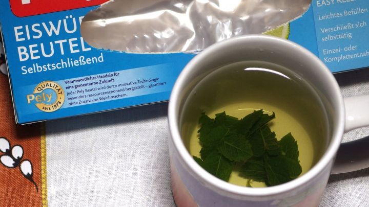 Eine Tasse Pfefferminztee mit frischen Bl&auml;ttern steht auf einem Tisch, umgeben von einem Eiw&uuml;rfel-Beutel.