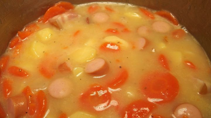 Ein Eintopf mit Karotten und W&uuml;rstchen kocht in einem Topf, wobei die Suppe leicht an den R&auml;ndern anbrannt ist.