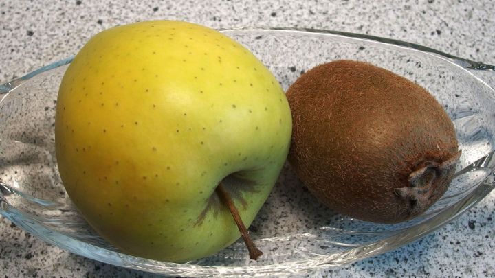 Ein gr&uuml;ner Apfel und eine Kiwi liegen auf einem Glasgericht, bereit zum Nachreifen in einer hellen K&uuml;chenumgebung.